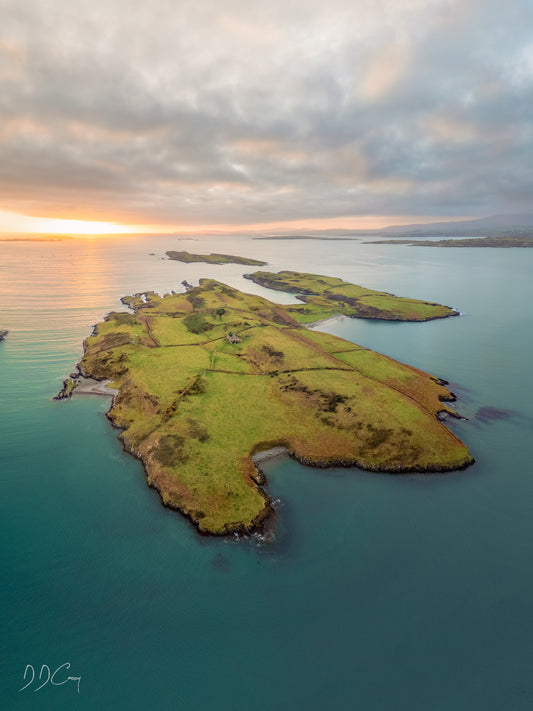 A sunset photo of Eastern Skeam Island in Ireland's Roaring Water Bay, with the outline of West Skeam Island and Brow Head in the distance. The island is surrounded by calm waters and the golden hour lighting adds warmth to the scene. The photo is a beautiful representation of the natural beauty of Ireland and a glimpse into its rich history.
