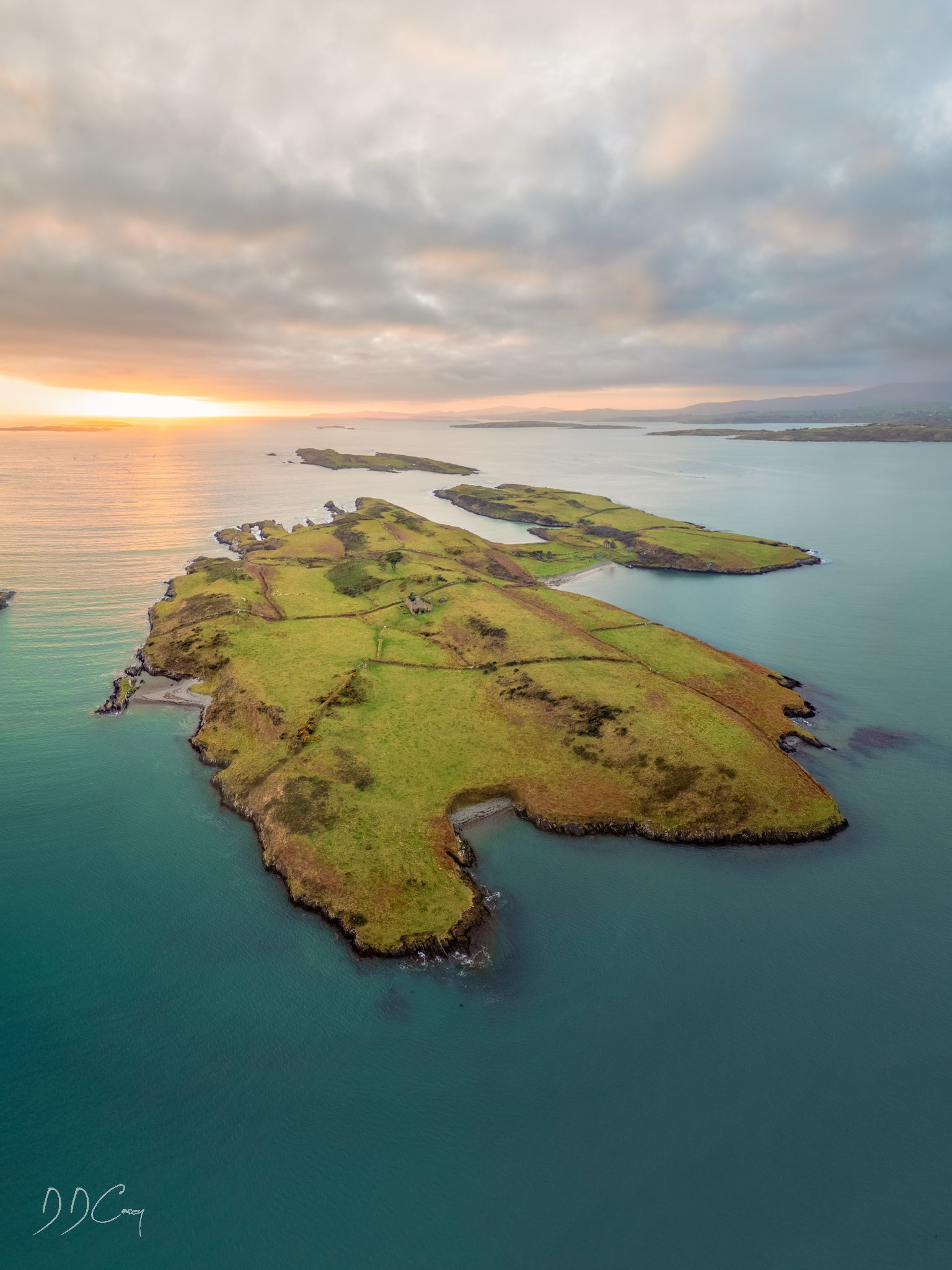 A sunset photo of Eastern Skeam Island in Ireland's Roaring Water Bay, with the outline of West Skeam Island and Brow Head in the distance. The island is surrounded by calm waters and the golden hour lighting adds warmth to the scene. The photo is a beautiful representation of the natural beauty of Ireland and a glimpse into its rich history.